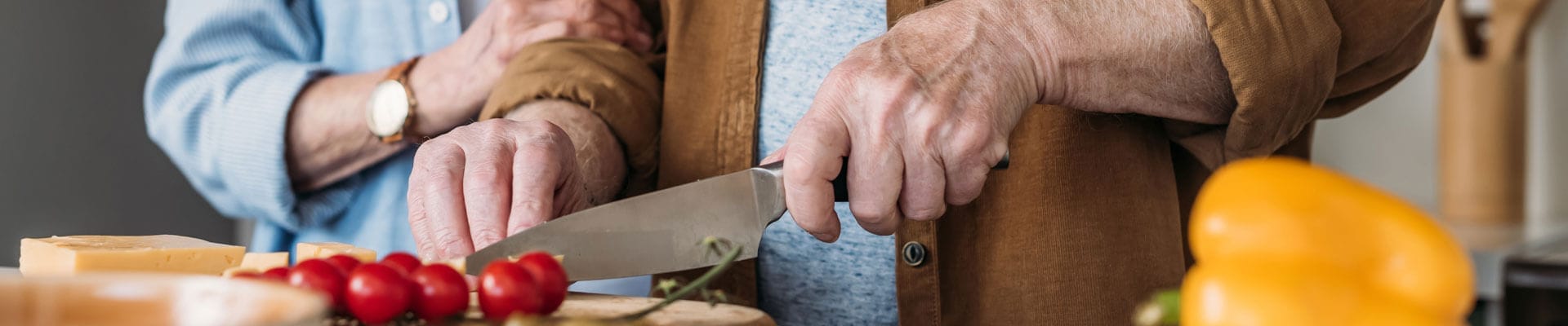cropped view of elderly woman near man cutting cheese on table with vegetables on blurred foreground, banner