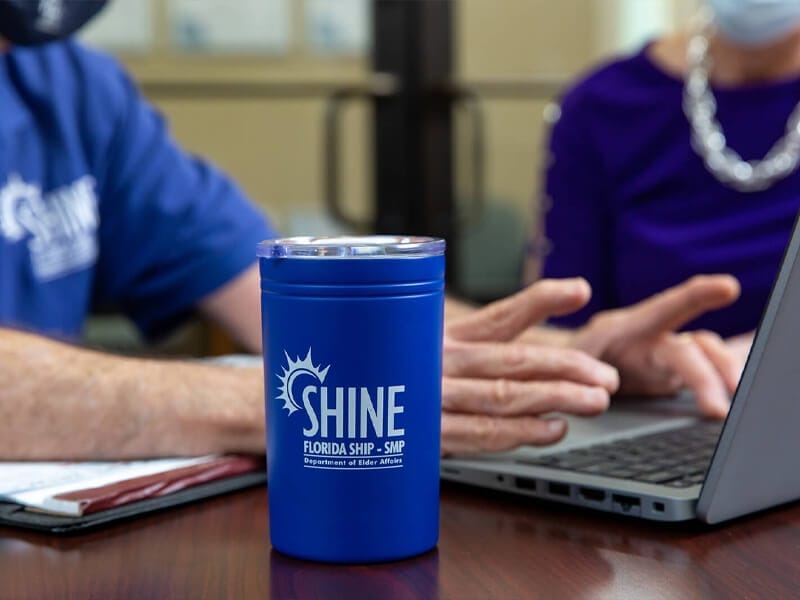 Image of a cup with the shine logo on it and a staff member working on a laptop in the background